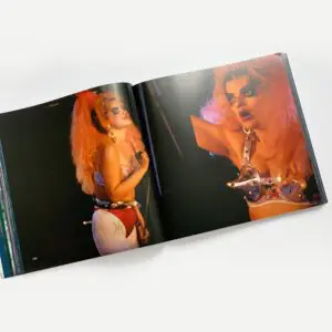 Un livre de photos s'ouvre sur une scène montrant une artiste aux grands cheveux orange, au visage maquillé et au soutien-gorge métallique clouté, posant sur scène sous un éclairage puissant.