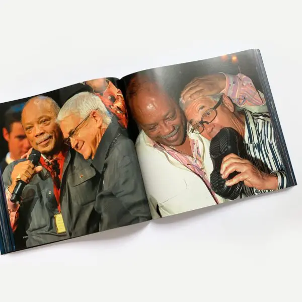 Livre ouvert présentant deux photos d'hommes âgés souriants. À gauche, deux hommes sont assis et parlent dans des microphones. À droite, deux hommes rient, l'un s'amusant à poser sa main sur la tête de l'autre tout en portant une chaussure à son oreille.