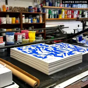 Une pile de tirages d'art avec des motifs abstraits bleus et blancs est posée sur une table de travail dans un studio d'impression, entourée de pots de peinture colorés et de fournitures. Une étiquette EDITION LIMITÉE apparaît dans le coin supérieur droit.