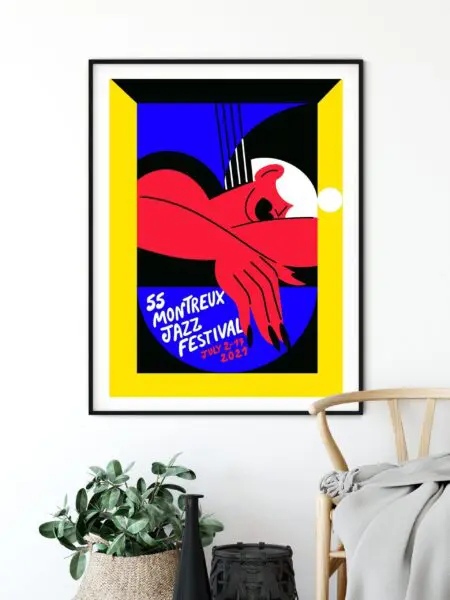 Affiche encadrée du Montreux Jazz Festival 2023 avec figure abstraite rouge, fond bleu et noir, bordure jaune, et texte indiquant 55 Montreux Jazz Festival 1-16 juillet 2023, affichée au-dessus d'une plante et d'une chaise.