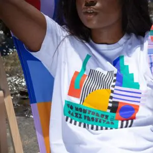 Une femme se détend à l'extérieur dans le T-Shirt Camille Walala, 2022, qui présente un graphique abstrait coloré et le texte "50 Montreux Jazz Festival". Une partie de son visage est visible alors qu'elle est assise sur une chaise multicolore.