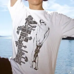 Une personne portant le T-Shirt Eric Wondergem, 1969 avec un graphique et un texte vintage du Festival de Jazz, se tenant à l'extérieur près d'un plan d'eau par une journée ensoleillée.