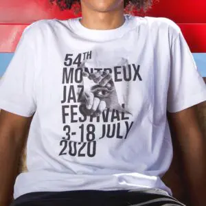 Une personne portant le T-Shirt JR, 2020 - un tee-shirt blanc portant le texte noir "54th Montreux Jazz Festival 3-18 July 2020" et un graphique d'œil regardant à travers une main sur le devant.