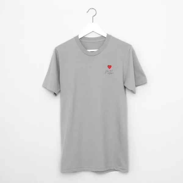 Un T-Shirt gris uni Cœur brodé, Montreux Jazz Festival, est suspendu à un cintre blanc contre un mur blanc, avec un petit cœur rouge et "Hope" en cursive sur la poitrine gauche.