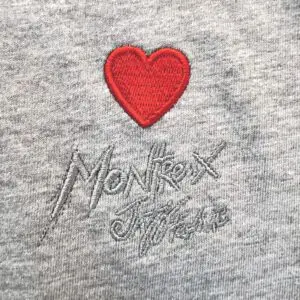 The T-Shirt Heart Embroidery, Montreux Jazz Festival features a red heart embroidered above gray "Montreux" stitching and a signature on soft heather gray fabric.