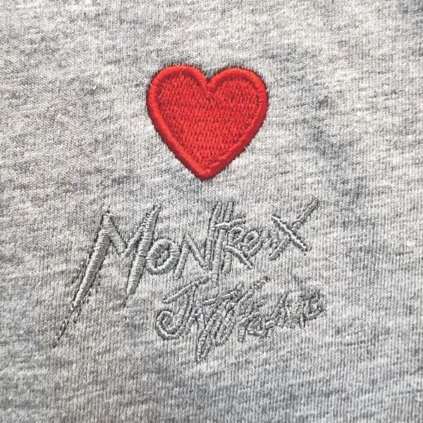 The T-Shirt Heart Embroidery, Montreux Jazz Festival features a red heart embroidered above gray "Montreux" stitching and a signature on soft heather gray fabric.