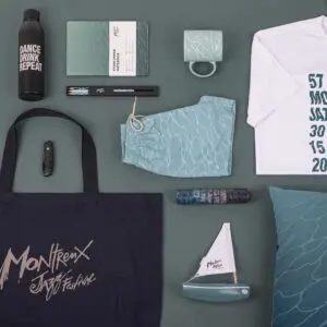 Vue à plat de produits dérivés du Festival de Jazz de Montreux, comprenant un fourre-tout, un T-shirt, un carnet de notes, une bouteille d'eau, un mug, un short, un stylo, un briquet, un oreiller, un étui à lunettes de soleil et un petit voilier décoratif, le tout dans des tons sarcelle et blanc.
