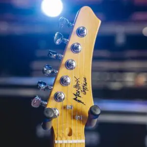 Gros plan d'une tête de guitare électrique avec des chevilles d'accord, des signatures et des lumières de scène floues en arrière-plan.