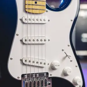 Gros plan d'une guitare électrique noire avec un pickguard blanc, trois micros, des boutons de réglage étiquetés volume et tonalité, et un manche en érable. L'arrière-plan est légèrement flou.