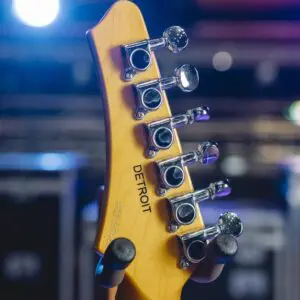 Gros plan sur l'arrière d'une tête de guitare avec des chevilles, le mot DETROIT gravé sur le bois, et des lumières de scène colorées floues en arrière-plan.