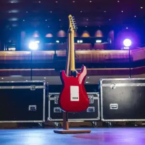 Une guitare électrique rouge sur pied est exposée au centre de la scène, le dos tourné vers la caméra. Des lumières de scène brillantes et de grandes caisses de matériel noires sont visibles à l'arrière-plan, suggérant un concert ou un spectacle.