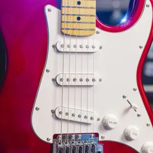 Gros plan d'une guitare électrique rouge avec une plaque de protection blanche, trois micros à simple bobinage, des boutons de commande et un manche en érable, montrant l'accastillage détaillé et la finition brillante.