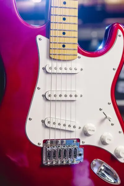 Gros plan d'une guitare électrique rouge avec une plaque de protection blanche, trois micros à simple bobinage, des boutons de commande et un manche en érable, montrant l'accastillage détaillé et la finition brillante.