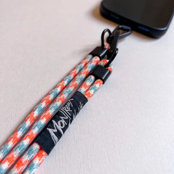 Close-up of a smartphone attached to the Mobile Strap, Lakeside—a colorful woven lanyard with orange, white, and blue stripes. It features a black "Mon Rope" tag and is secured to the case with a black metal clasp.