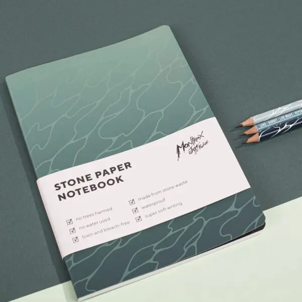 A green Stone Paper Notebook, Guillaume “SupaKitch,” with a wavy cover sits on a green surface by two pencils. The label notes it’s made from stone waste, waterproof, offers super soft writing, and uses no trees.