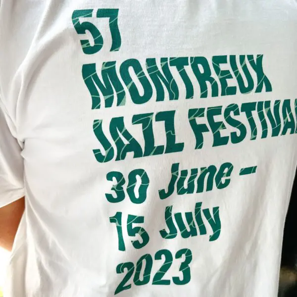 Gros plan sur un T-shirt blanc avec un texte vert indiquant "57 Montreux Jazz Festival 30 juin - 15 juillet 2023". Le T-shirt est porté par une personne dont on ne voit qu'une partie du dos et du bras.