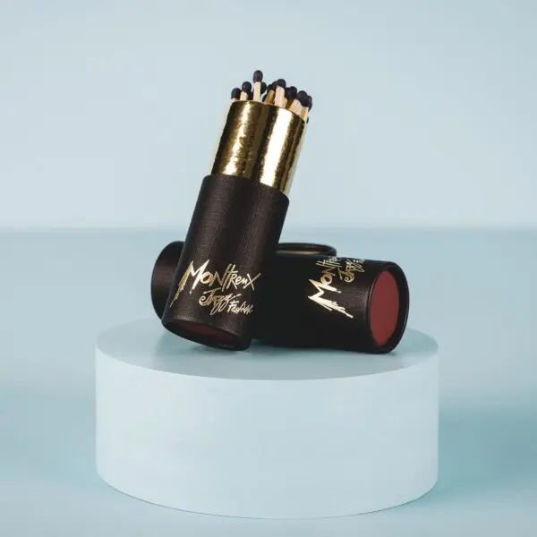 Two black and gold cylindrical containers labeled "Matchbox" hold dark-tipped matches, displayed on a light blue round platform against a matching background—evoking the timeless style of the Montreux Jazz Festival.