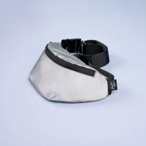 The Reflective Mini Bumbag in light gray, featuring a black zipper, adjustable strap, and small black tag, is displayed on a smooth light blue surface.