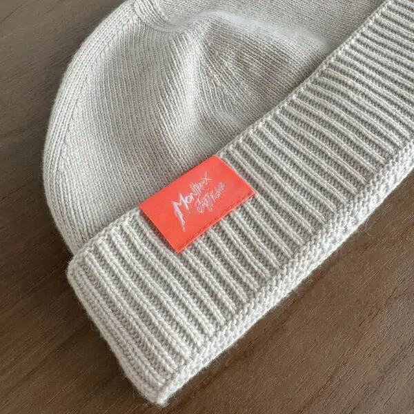 Un bonnet en tricot beige avec un large bord plié, posé sur une surface en bois. Le bonnet comporte une étiquette orange avec un texte et des signatures en blanc.