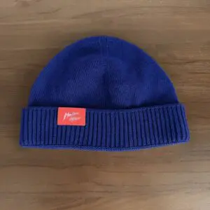 Un bonnet en tricot bleu avec un bord plié et un écusson rectangulaire rouge comportant un texte manuscrit blanc, placé sur une surface en bois.