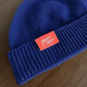 Un bonnet en tricot bleu avec un bord replié, comportant un écusson rouge vif avec un texte écrit à la main en blanc indiquant Montreux Jazz Festival. Le bonnet est posé sur une surface en bois.