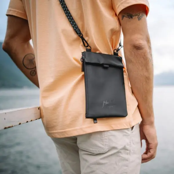 A person wearing a peach t-shirt and light shorts stands by the water at the Montreux Festival, carrying the Festival Bag, Lakeside with a visible logo. Mountains and a cloudy sky complete the scenic outdoor background.