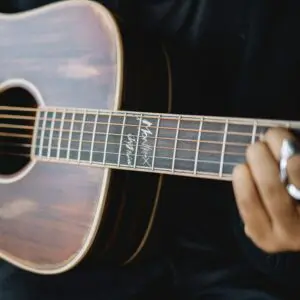 Gros plan d'une personne jouant d'une guitare acoustique, se concentrant sur sa main appuyant sur les cordes du manche. Certaines signatures ou dessins sont visibles sur le manche, près du corps de la guitare.