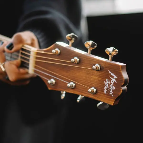 Gros plan sur la main d'une personne tenant le manche d'une guitare acoustique, en mettant l'accent sur la tête de la guitare, les chevilles d'accordage et le logo. La personne a les ongles vernis en noir et porte des bagues.