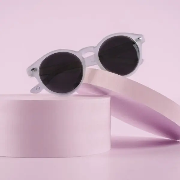 Fly Bruxelles Cristal Glasses, featuring white frames and round black lenses inspired by Montreux Jazz style, are showcased on pastel pink cylindrical, slanted platforms against a minimalist pink background.
