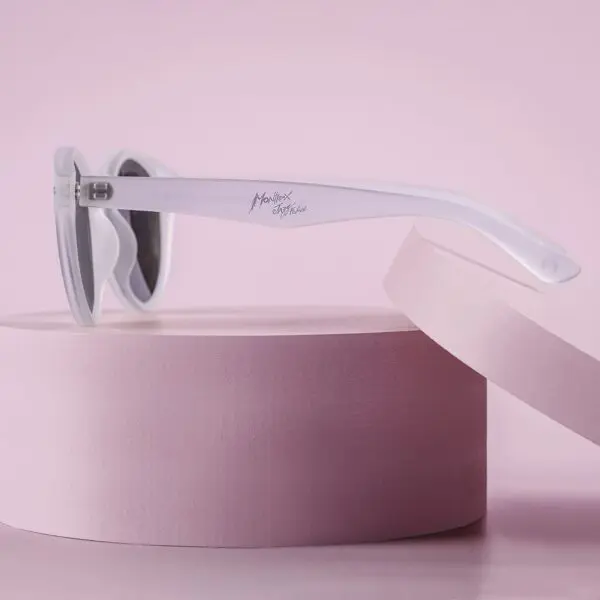 A pair of Fly Bruxelles Cristal Glasses with gray lenses rests on a pink round platform, accompanied by another pink circular object, all set against a pink background.