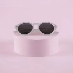 Fly Bruxelles Cristal Glasses, white round sunglasses with dark lenses, rest on a cylindrical pink pedestal against a pastel pink background.