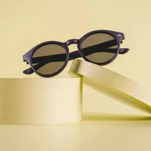 Les lunettes de soleil rondes Lunettes Fly Bruxelles Noir Mat aux verres foncés reposent élégamment sur deux supports cylindriques jaune pâle sur un fond jaune assorti.