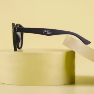 The Fly Bruxelles Matte Black Glasses with thick frames are displayed on a round, light yellow pedestal against a matching background. The temple arm features a white Montreux Jazz Festival logo.