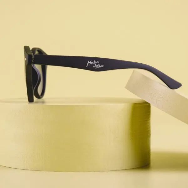 The Fly Bruxelles Matte Black Glasses with thick frames are displayed on a round, light yellow pedestal against a matching background. The temple arm features a white Montreux Jazz Festival logo.