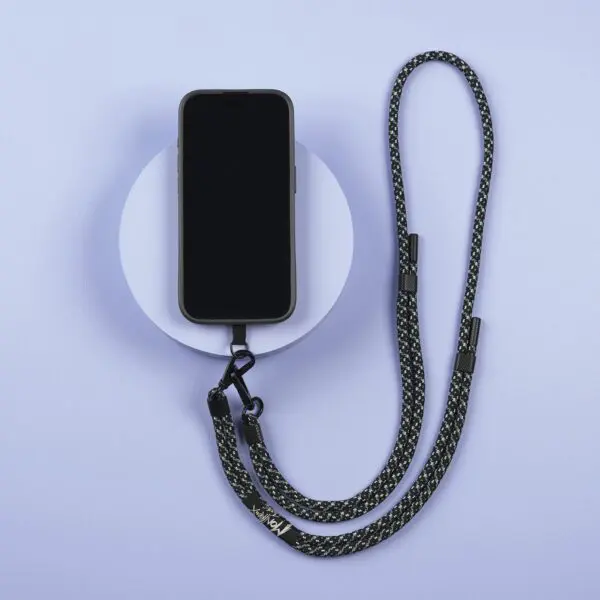 A smartphone with a black case is attached to a Mobile Strap, Lakeside and placed on a light purple background with a circular white platform underneath.