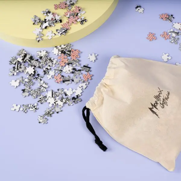 Un sac à cordon beige avec un texte noir est posé sur une surface violette claire à côté de pièces de puzzle éparpillées, certaines en noir et blanc, d'autres en rose. Un objet courbe jaune pâle apparaît au sommet.