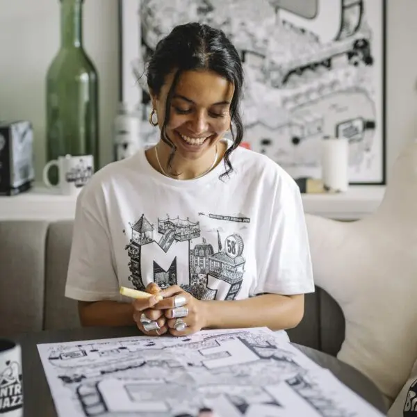 Une femme aux cheveux noirs bouclés, vêtue d'un T-shirt graphique blanc, sourit tout en dessinant avec un marqueur à une table couverte d'œuvres d'art en noir et blanc. Des fournitures artistiques et des illustrations détaillées sont visibles autour d'elle.