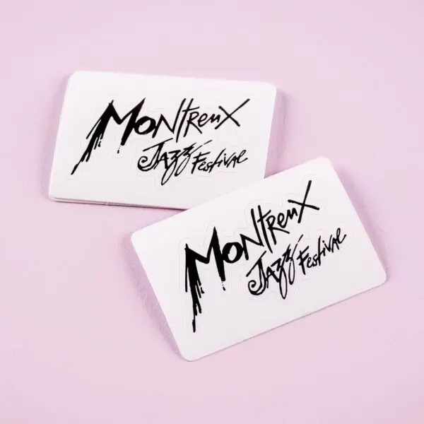 Deux cartes Sticker blanc Montreux Jazz Festival, arborant le logo noir du festival, se chevauchent légèrement sur une surface rose pastel.