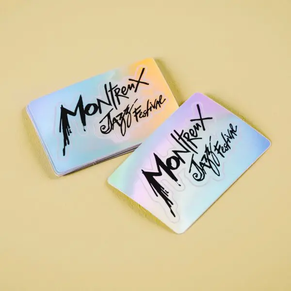 Deux cartes Sticker holographique Montreux Jazz Festival avec une finition brillante, irisée par l'arc-en-ciel et une écriture noire audacieuse sont placées sur une surface beige clair.
