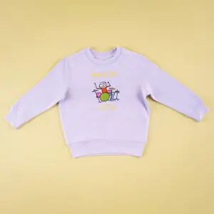 Pull à manches longues Phil Collins, Enfant, de couleur mauve clair, avec une caricature colorée d'une personne souriante jouant de la batterie et le texte "Midweek Jam, July 3rd 1989" sur le devant, sur fond beige.