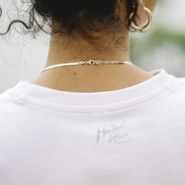 Gros plan de dos sur le cou et les épaules d'une personne, montrant un T-shirt blanc avec un léger logo "Montreux Jazz Festival" imprimé au dos, un fin collier, des cheveux bouclés et une boucle d'oreille en or.