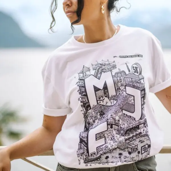 Une femme se tient à l'extérieur, près d'une balustrade, et porte le T-shirt 2024 Rylsee, avec un lettrage MFJ audacieux et un graphisme noir complexe, célébrant le festival de Montreux. L'eau, les montagnes et la verdure apparaissent floues en arrière-plan.