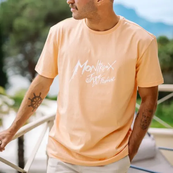 Vêtue d'un T-Shirt Montreux Jazz Festival, Pêche, une personne s'appuie sur une balustrade en plein air, avec en arrière-plan des arbres et des montagnes aux contours flous.