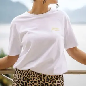 MJF24-T-shirt-Transfert-Jaune-Montreux-Jazz-Festival-Retouche-1.jpg Une personne portant un T-Shirt blanc Montreux Jazz, Butter avec un petit texte doré sur la poitrine et un pantalon imprimé léopard se tient à l'extérieur, appuyée sur une balustrade avec un arrière-plan naturel flou.