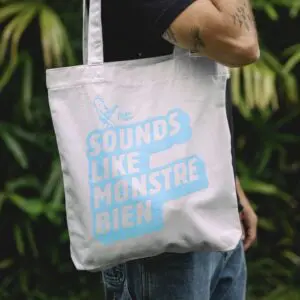 Une personne tient le Tote Bag Rylsee, Bleu, avec un texte bleu clair "SOUNDS LIKE MONSTRE BIEN" et un monstre jouant de la guitare. En arrière-plan, un feuillage vert encadre le motif inspiré de Montreux Bleu.