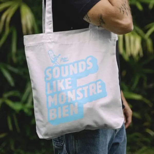 A person holds the Tote Bag Rylsee, Blue, featuring light blue "SOUNDS LIKE MONSTRE BIEN" text and a guitar-playing monster. Lush green plants are blurred in the background, creating a fresh Montreux Blue ambiance.