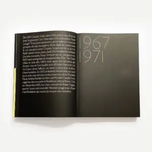 Un livre ouvert présente une page noire avec un texte en français à gauche et de grands chiffres blancs et or 1967 1971 à droite, le tout sur un fond blanc minimaliste.