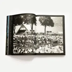 Livre ouvert présentant une photographie en noir et blanc d'un grand groupe de personnes assises sur l'herbe, face à une scène installée au bord d'un lac, avec des arbres et des montagnes en arrière-plan.