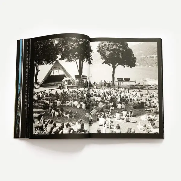 Livre ouvert présentant une photographie en noir et blanc d'un grand groupe de personnes assises sur l'herbe, face à une scène installée au bord d'un lac, avec des arbres et des montagnes en arrière-plan.