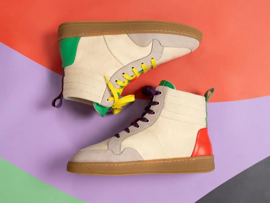 Deux baskets montantes en daim beige, avec des accents verts et rouges sur les talons, des lacets jaunes et violets, et des semelles marron, sont présentées sur un fond abstrait coloré avec des formes rouges, violettes et vertes.
